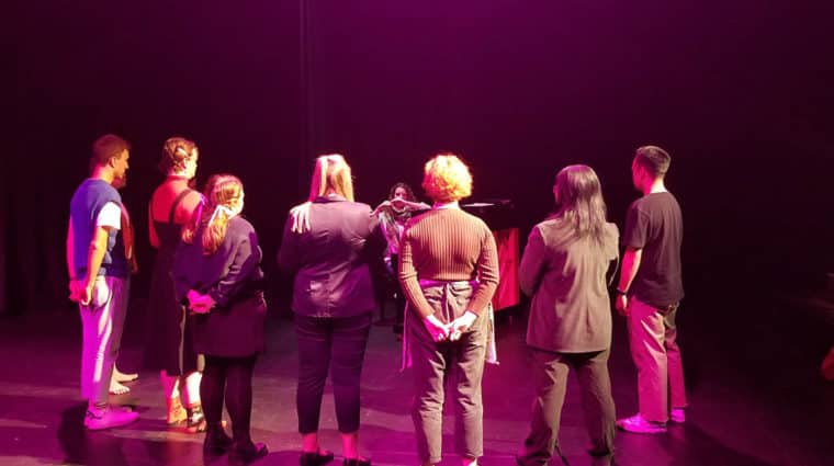 Students standing in a semi-circle on a stage looking towards a person at a piano talking to them.